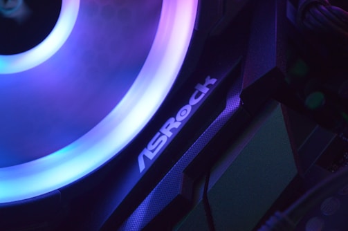 A close-up view of a computer component with a glowing LED ring in shades of blue and purple. The logo 'ASRock' is visible on the device, highlighting part of the internal hardware setup.
