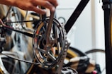 A close-up of a bicycle gear and chain with a person's hand working on it. The focus is on the metal components, showing precise details of the gear and chain mechanism. The background includes blurred out parts of the bicycle and other equipment.