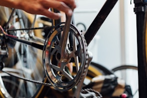 A close-up of a bicycle gear and chain with a person's hand working on it. The focus is on the metal components, showing precise details of the gear and chain mechanism. The background includes blurred out parts of the bicycle and other equipment.