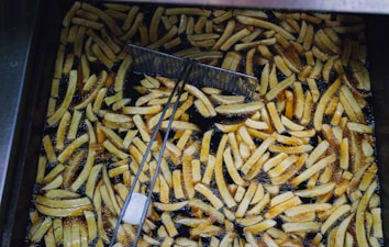 A large batch of golden-brown French fries is frying in hot oil within a deep fryer. Numerous fries are visible, some fully submerged and others partially above the oil. A metal wire mesh scoop is positioned in the fryer, likely used for stirring or removing the fries.