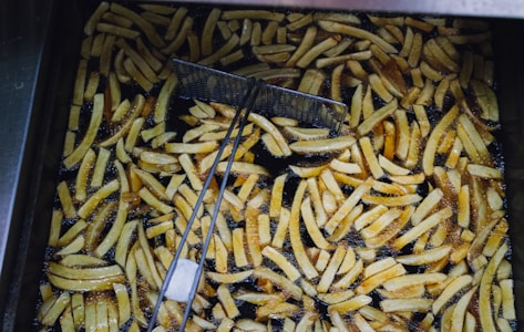A large batch of golden-brown French fries is frying in hot oil within a deep fryer. Numerous fries are visible, some fully submerged and others partially above the oil. A metal wire mesh scoop is positioned in the fryer, likely used for stirring or removing the fries.