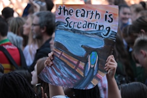 A crowd of people gathered, with someone holding up a protest sign that reads 'the earth is screaming'. The sign features an illustration inspired by Edvard Munch's 'The Scream', depicting the Earth in place of the face, conveying an environmental message.