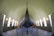 A large, ancient Viking ship is displayed in a museum setting with high arched ceilings. The ship's dark wooden hull stands prominently in the center, surrounded by visitor barriers. Natural light floods in through tall, narrow windows along the walls, highlighting the detailed craftsmanship of the ship. Several people are observing the exhibit, adding a sense of scale to the impressive structure.