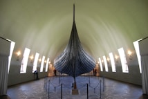 A large, ancient Viking ship is displayed in a museum setting with high arched ceilings. The ship's dark wooden hull stands prominently in the center, surrounded by visitor barriers. Natural light floods in through tall, narrow windows along the walls, highlighting the detailed craftsmanship of the ship. Several people are observing the exhibit, adding a sense of scale to the impressive structure.