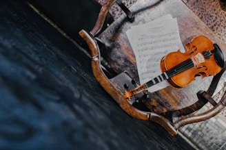 A wooden chair with a violin and sheet music placed on its seat. The violin has a warm, polished brown finish and is resting on top of the sheet music. The chair is situated on a dark, textured floor, adding contrast to the scene.