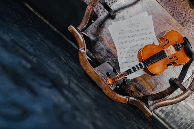 A wooden chair with a violin and sheet music placed on its seat. The violin has a warm, polished brown finish and is resting on top of the sheet music. The chair is situated on a dark, textured floor, adding contrast to the scene.