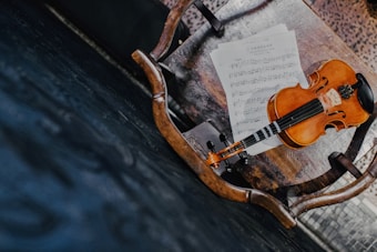 A wooden chair with a violin and sheet music placed on its seat. The violin has a warm, polished brown finish and is resting on top of the sheet music. The chair is situated on a dark, textured floor, adding contrast to the scene.