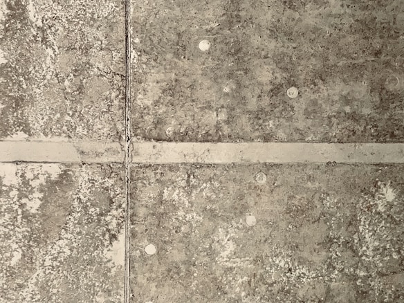 A close-up view of a textured concrete surface with visible lines and patches of wear. The surface has a grid pattern formed by intersecting lines, which are likely seams or joints in the concrete panels. The texture is uneven, with spots of discoloration and small, circular marks that appear consistently across the surface.