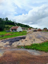 A solitary small white house with a red tiled roof sits on a rocky landscape surrounded by lush green grass and hills. Several haystacks are scattered around, under a dynamic cloudy sky.