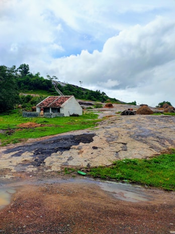 A solitary small white house with a red tiled roof sits on a rocky landscape surrounded by lush green grass and hills. Several haystacks are scattered around, under a dynamic cloudy sky.