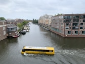 A yellow amphibious tour bus is traveling along a narrow canal lined with brick buildings and greenery. The architecture is modern with large windows, and several smaller boats are docked along the canal. The sky is overcast, adding a calm and serene atmosphere to the scene.