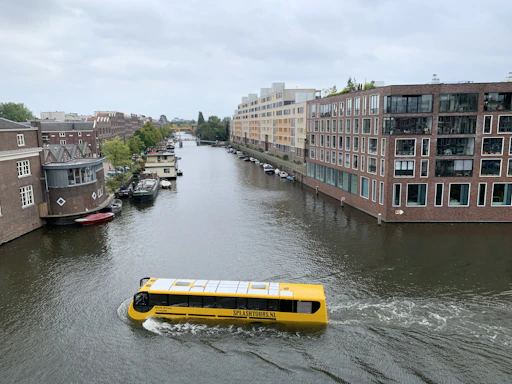 A yellow amphibious tour bus is traveling along a narrow canal lined with brick buildings and greenery. The architecture is modern with large windows, and several smaller boats are docked along the canal. The sky is overcast, adding a calm and serene atmosphere to the scene.