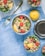 Bowls of salad containing quinoa, chickpeas, tomatoes, cucumbers, olives, and lemon wedges are arranged on a blue cloth. A fork and a whole lemon are also seen on the cloth, along with a metal container lid.