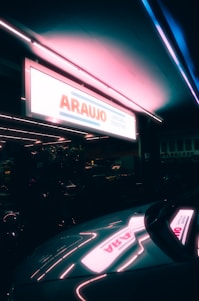A glowing neon sign for a drugstore with the name 'ARAUJO' is prominently displayed. The sign casts a colorful reflection on the surface of a parked car nearby, creating a vibrant and moody atmosphere. The surroundings are dimly lit, suggesting a night-time or twilight setting.