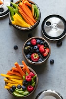 Three small containers filled with an assortment of fresh vegetables and fruits, including sliced cucumbers, carrots, yellow bell peppers, snap peas, strawberries, blackberries, blueberries, and raspberries. The containers are circular and have removable black and silver lids, with a few berries scattered around them on a light gray surface.