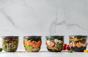 Four containers filled with assorted, colorful salads are placed in a row against a light marble background. Each container has a unique combination of vibrant vegetables, grains, and toppings, showcasing a variety of textures and layers.