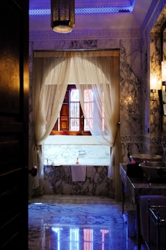 A luxurious bathroom adorned with intricate marble patterns on the floor and walls, complemented by a large wooden-framed window draped with elegant sheer curtains. The ambient lighting includes a central hanging lantern with ornate detailing, casting soft glows. A well-appointed bathtub rests beneath the window, accompanied by a neatly folded towel and a bottle.
