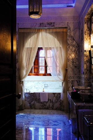 A luxurious bathroom adorned with intricate marble patterns on the floor and walls, complemented by a large wooden-framed window draped with elegant sheer curtains. The ambient lighting includes a central hanging lantern with ornate detailing, casting soft glows. A well-appointed bathtub rests beneath the window, accompanied by a neatly folded towel and a bottle.