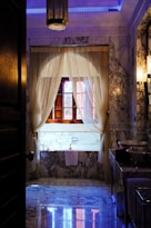 A luxurious bathroom adorned with intricate marble patterns on the floor and walls, complemented by a large wooden-framed window draped with elegant sheer curtains. The ambient lighting includes a central hanging lantern with ornate detailing, casting soft glows. A well-appointed bathtub rests beneath the window, accompanied by a neatly folded towel and a bottle.