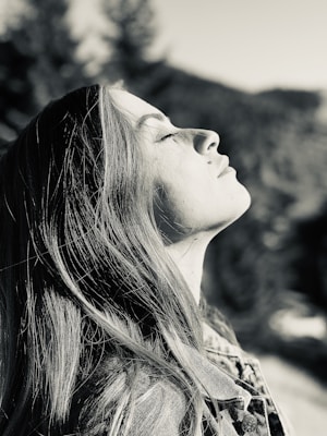 A person with long hair is facing upwards with closed eyes, appearing relaxed and at peace. The image is in black and white, adding a timeless quality. Soft-focus background suggests an outdoor setting with trees.