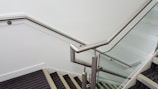 A modern staircase with a sleek metal handrail and glass panels along the sides. The steps are covered with a dark carpet, and the walls are painted white.