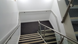 A staircase with a modern design features glass panels and metallic railings. The steps are covered with a dark carpet in a pattern, and the walls are painted white, creating a clean and minimalist appearance.