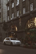 A silver luxury SUV is parked on a city street in front of a Gucci store. The building facade is ornate with large arched windows and decorative elements, including sculptures. The scene is urban, with greenery on the sidewalk and warm lights illuminating the storefront.