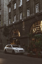 A silver luxury SUV is parked on a city street in front of a Gucci store. The building facade is ornate with large arched windows and decorative elements, including sculptures. The scene is urban, with greenery on the sidewalk and warm lights illuminating the storefront.