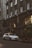 A silver luxury SUV is parked on a city street in front of a Gucci store. The building facade is ornate with large arched windows and decorative elements, including sculptures. The scene is urban, with greenery on the sidewalk and warm lights illuminating the storefront.