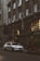 A silver luxury SUV is parked on a city street in front of a Gucci store. The building facade is ornate with large arched windows and decorative elements, including sculptures. The scene is urban, with greenery on the sidewalk and warm lights illuminating the storefront.