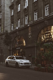A silver luxury SUV is parked on a city street in front of a Gucci store. The building facade is ornate with large arched windows and decorative elements, including sculptures. The scene is urban, with greenery on the sidewalk and warm lights illuminating the storefront.