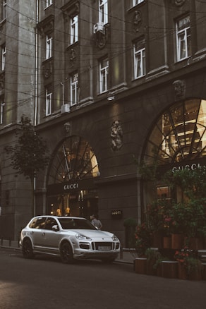 A silver luxury SUV is parked on a city street in front of a Gucci store. The building facade is ornate with large arched windows and decorative elements, including sculptures. The scene is urban, with greenery on the sidewalk and warm lights illuminating the storefront.