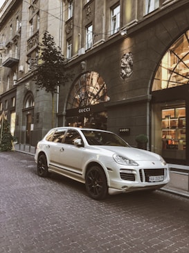 A luxury SUV is parked on a cobblestone street in front of a Gucci store located in an elegant building. The architecture features ornate details and large arched windows, reflecting a sense of sophistication. Small trees and decorative lighting add to the upscale urban setting.