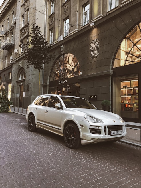 A luxury SUV is parked on a cobblestone street in front of a Gucci store located in an elegant building. The architecture features ornate details and large arched windows, reflecting a sense of sophistication. Small trees and decorative lighting add to the upscale urban setting.