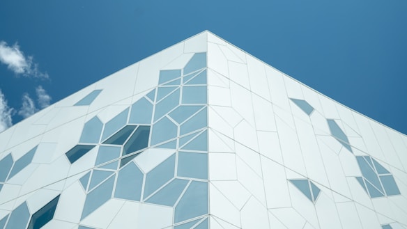 A modern building facade featuring geometric patterns of angular shapes and windows. The structure is set against a clear blue sky with a few scattered clouds, giving the design a clean and contemporary appearance. The use of light colors enhances the crisp, minimalist aesthetic.