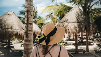 A person with long hair is wearing a wide-brimmed straw hat adorned with a black ribbon, standing on a tropical beach. Thatched-roof umbrellas made of palm leaves are surrounded by tall palm trees. Sunloungers are scattered on the sand under the shade of the umbrellas, creating a serene and relaxing atmosphere.