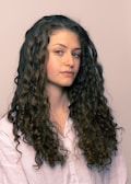 A person with long, curly brown hair wearing a light-colored shirt is looking slightly to the side. The hair is voluminous and well-defined, cascading down the shoulders.