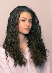 A person with long, curly brown hair wearing a light-colored shirt is looking slightly to the side. The hair is voluminous and well-defined, cascading down the shoulders.