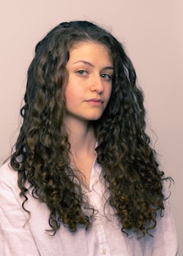 A person with long, curly brown hair wearing a light-colored shirt is looking slightly to the side. The hair is voluminous and well-defined, cascading down the shoulders.