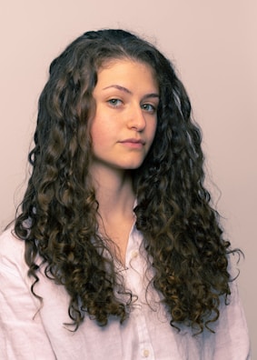 A person with long, curly brown hair wearing a light-colored shirt is looking slightly to the side. The hair is voluminous and well-defined, cascading down the shoulders.