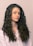 A person with long, curly brown hair wearing a light-colored shirt is looking slightly to the side. The hair is voluminous and well-defined, cascading down the shoulders.