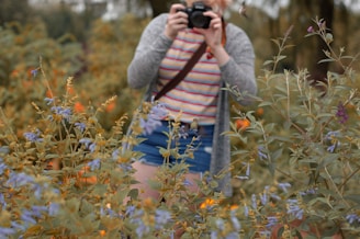 A person dressed casually in a striped shirt and shorts is taking a photograph in a lush garden. They are surrounded by abundant foliage and various flowers, creating an impression of being deeply immersed in nature.