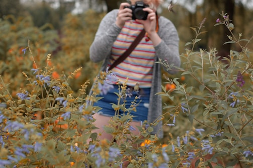 A person dressed casually in a striped shirt and shorts is taking a photograph in a lush garden. They are surrounded by abundant foliage and various flowers, creating an impression of being deeply immersed in nature.