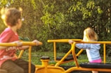 Two children are playing on a merry-go-round in a park surrounded by lush green foliage. One child is in a red shirt and the other in a striped hoodie, both holding onto the yellow metal bars of the playground equipment. The setting is bright with natural sunlight filtering through the trees, creating a warm and lively atmosphere.