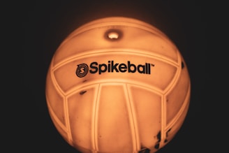 A close-up view of a ball with the logo 'Spikeball' visible on its surface. The ball appears to be illuminated with a warm, yellow-orange glow, accentuating its texture and design.