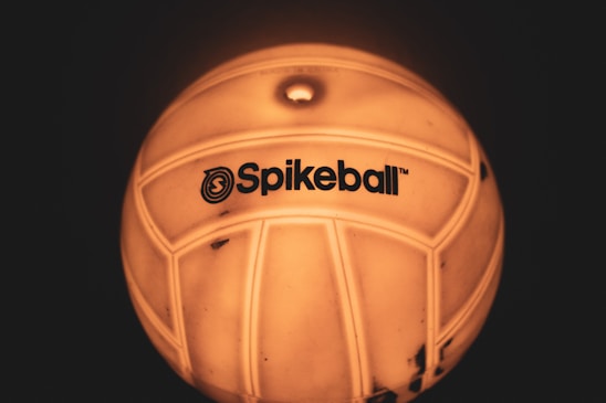 A close-up view of a ball with the logo 'Spikeball' visible on its surface. The ball appears to be illuminated with a warm, yellow-orange glow, accentuating its texture and design.