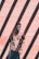 A man wearing sunglasses and a colorful Hawaiian shirt stands against a salmon-pink tiled wall. Diagonal shadows cast by a structure above create stripes across the wall and the man.