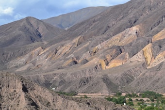 A rugged mountain landscape with vibrant geological formations featuring layers of earthy browns, oranges, and yellows. In the foreground, sparse vegetation and a small patch of greenery can be seen at the base of the mountains.