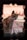 A cat with a mix of white, brown, and black fur is framed by a decorative hole in a wall. The animal sits on a colorful, patterned tile floor with a warm, pinkish wall as the backdrop.