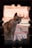 A cat with a mix of white, brown, and black fur is framed by a decorative hole in a wall. The animal sits on a colorful, patterned tile floor with a warm, pinkish wall as the backdrop.